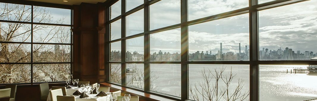 Dining tables beside tall windows overlooking the Hudson River at Fleming’s Edgewater.